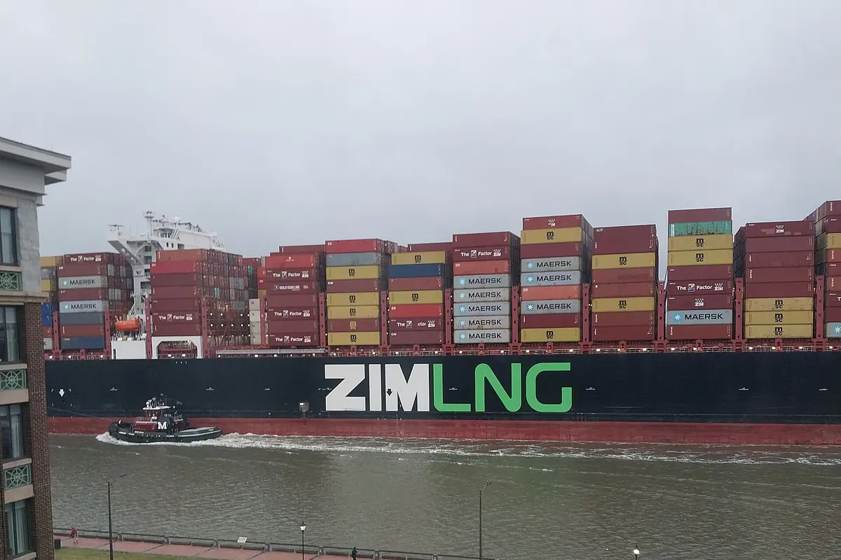 A loaded container ship passing the Savannah riverfront, with a small tug beside it.