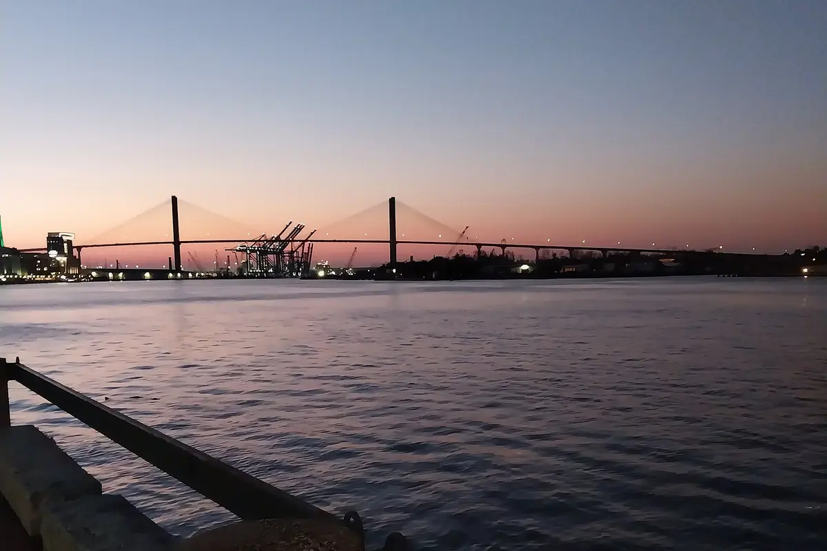 The Talmadge Memorial Bridge and the port cranes in silhouette against a pink and violet dusk sky over the Savannah River.
