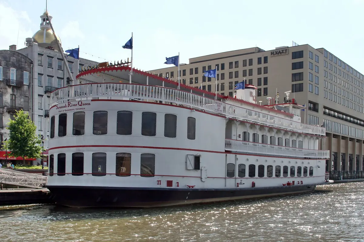 The white paddlewheel riverboat Savannah River Queen at its berth beside River Street, with a gold dome and hotel buildings behind it.
